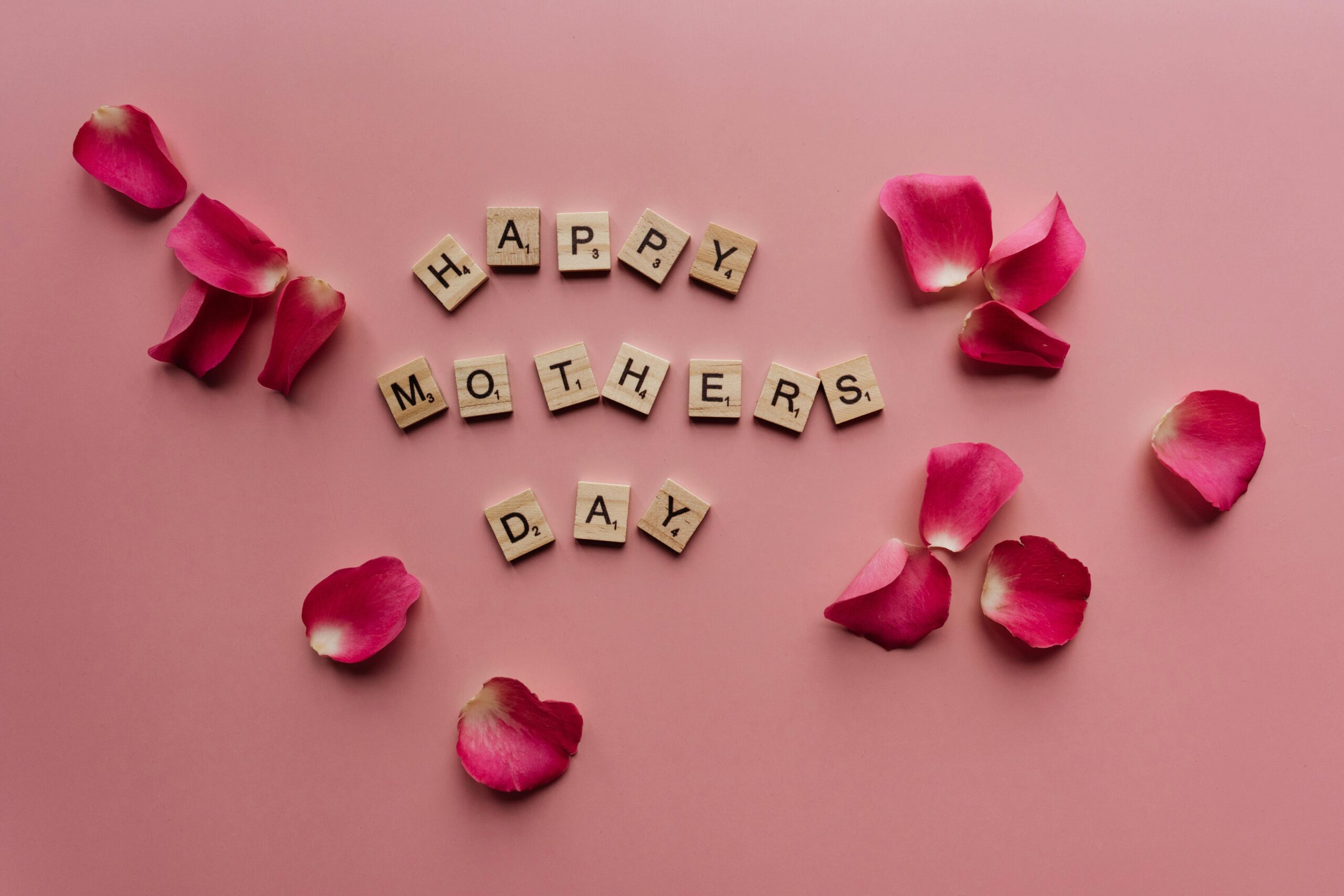 Wooden Scrabble tiles spelling “Happy Mother’s Day” surrounded by pink rose petals on a soft pink background.