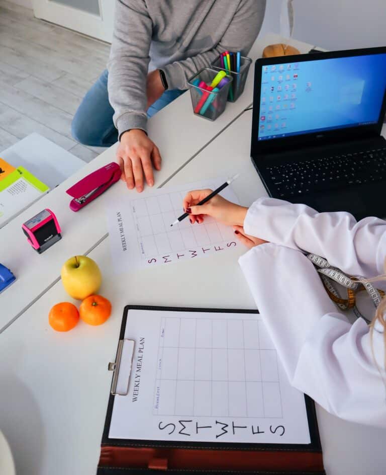 Two people planning a weekly meal schedule at a bright workspace with fruits, stationery, and a laptop nearby.